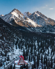 A red-roofed mountain lodge surrounded by snow-dusted pine trees set against a backdrop of rugged, sunlit, snow-capped mountains and a blue sky