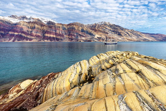 The amazing colourful rock formations of Segelsalskapet fjord, in the Northeast Greenland National Park. A tall sailing ship and the Berzelius Bjerg Mountain can be seen behind.