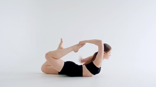 Girl holding bow pose by bending her back and grasping feet firmly while lying on stomach during hatha yoga practice in minimalist studio with white background