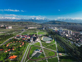 Wide aerial view of a large, green urban park area and residential buildings set against the dramatic, snow-capped Tian Shan mountains