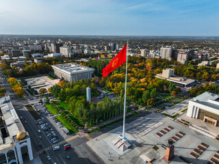 Aerial view of the large red Kyrgyz national flag on a flagpole over Ala-Too Square, surrounded by city buildings and autumn trees