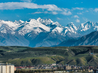 Telephoto shot of the jagged, snow-covered Tian Shan peaks rising dramatically above rolling green foothills and apartment buildings