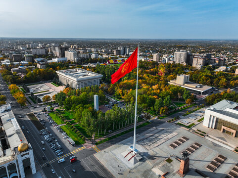 Aerial view of the large red Kyrgyz national flag on a flagpole over Ala-Too Square, surrounded by city buildings and autumn trees