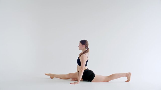 Girl performing sequence of yoga poses, such as one legged downward facing dog, full front split and center split on floor in white minimalist studio