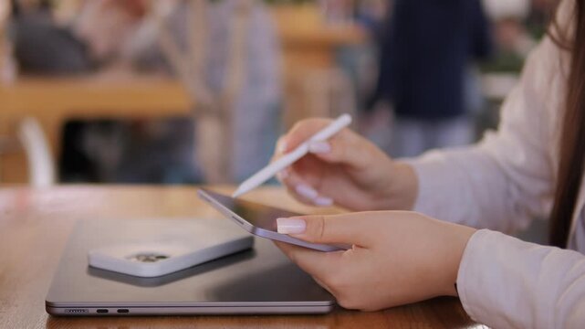 Woman is holding a pen and a tablet. She is sitting at a table. There are other people in the background