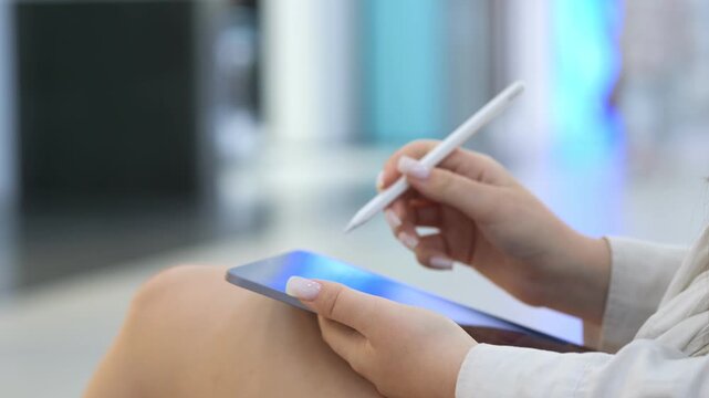 Woman is holding a tablet and a pen. She is sitting down. There are other people in the background