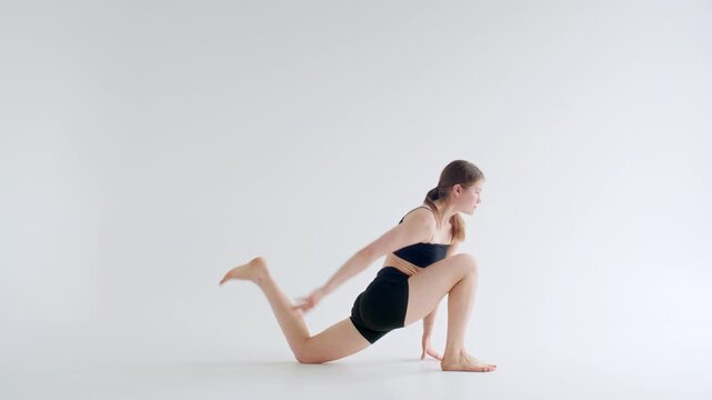 Young woman in sports bra and shorts performing downward facing dog pose, quad stretch and low lunge backbend variation with foot close to her head during yoga practice in minimalist white studio