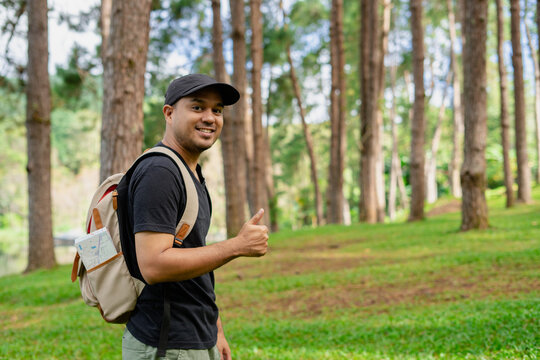 Young happy man standing and amile showing thumb up enjoying the fresh air in the forest. Male hiker with backpack and map with open arms enjoying and relax the nature.