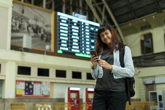Traveler female at train station check-in on smartphone  railway platform. Asian woman with timetables information of departures arrivals on behind. Tourist adventure holiday traveling concept.