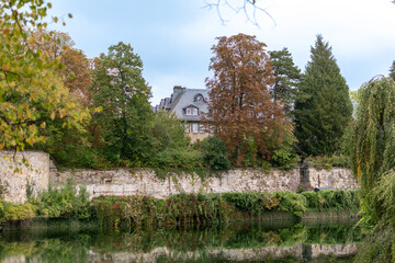 Stately house nestled behind an old, long stone wall and surrounded by trees displaying a mix of green and autumnal brown and yellow foliage, reflected in the water below