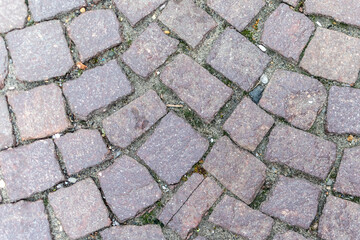 Top-down, close-up photograph of a cobblestone street or pavement, featuring reddish-brown, roughly rectangular stones set in an irregular pattern with visible dirt and grit in the joints