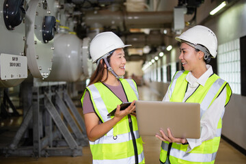 ฟCommunicating and sharing ideas at a high voltage substation, two female engineers use a laptop and tablet to discuss safety, coordinate inspections, and manage energy operations with teamwork