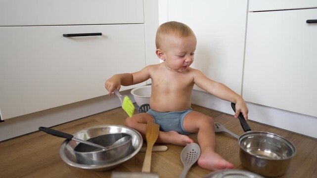 Baby play with kitchen utensil toddler sits on floor near cabinet holding pot lid and spoon exploring pan with teether and spoon creating mess in kitchen while child curious and innocent and