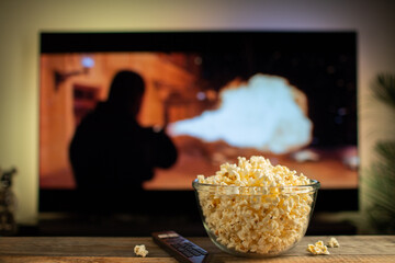 A glass bowl of popcorn and remote control in the background the TV works. Evening cozy watching a...
