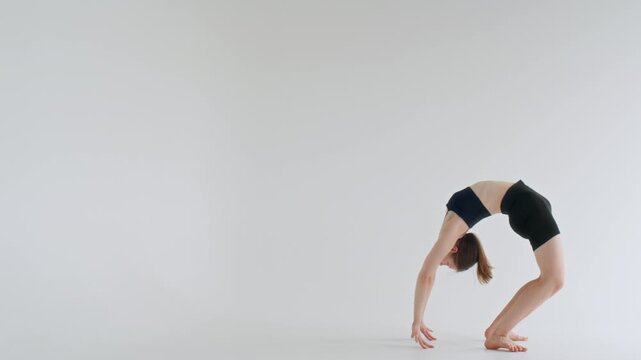 Young flexible woman in sports bra and shorts arching her back and lifting one leg high while performing one-legged wheel pose in minimalist white studio with copy space
