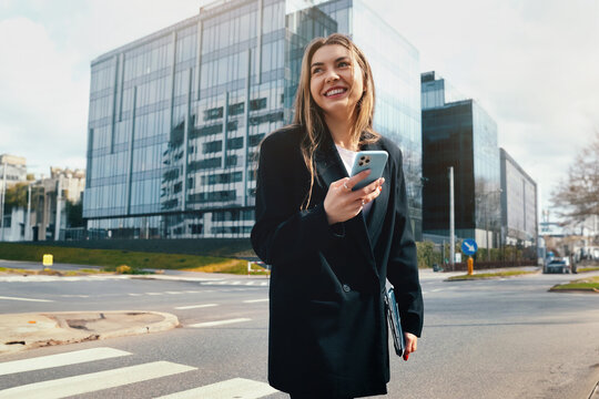 Woman in Stylish Outfit Walking Confidently on City Street While Using Smartphone Near Modern Buildings During Daytime