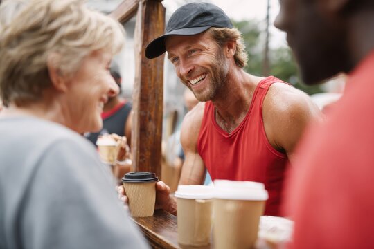 A cheerful, athletic man in a red tank top receives coffee from a friendly senior couple at a race stall, showcasing community spirit and engagement during a lively outdoor event.