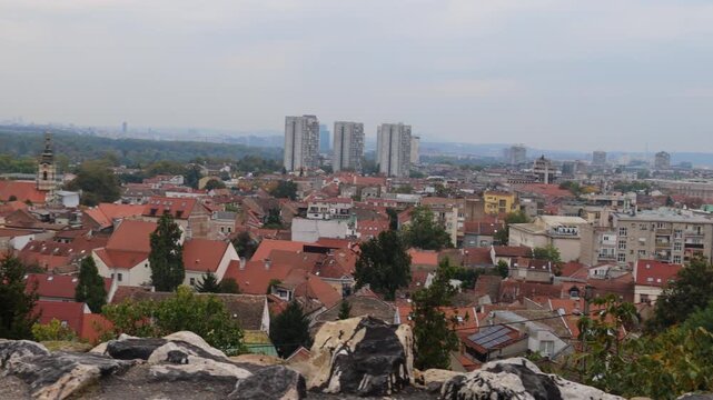A scenic view of Zemun, a historic district of Belgrade, Serbia, featuring red-tiled rooftops, old houses, and church towers by the Danube River. In the distance, modern New Belgrade buildings rise