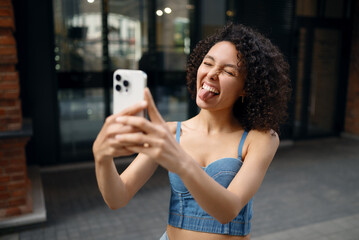 Playful young woman taking a funny selfie with her smartphone outdoors, sticking out tongue and laughing. Concept for fun, self-expression, and social media lifestyle.