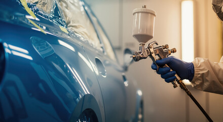 A professional painter sprays a blue car with a spray gun in an auto body shop. Automotive vehicle restoration and repair service. Close-up on the painting process