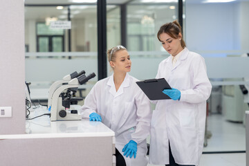 Fototapeta premium Two women scientists collaborate in a modern laboratory reviewing research data while using a microscope
