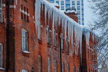 Sharp icicles hanging from weathered brick roof, winter landscape contrasting modern architecture...