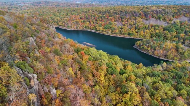 Aerial drone shot of Merimere Reservoir Loop in Meriden, Connecticut. Colorful fall foliage, rugged cliffs, winding lake, and expansive forest offer tranquil beauty for outdoor and nature themes.