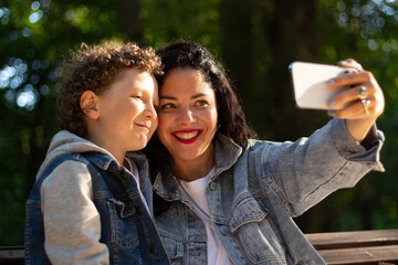 Young mother making selfie or video call with her son sitting on bench in park. Mom and boy calling grandparents. Concept of: connection, distance, technology