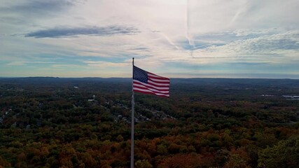 Aerial shot of an American flag waving above autumn forest and distant hills under a dramatic, partly cloudy sky. Castle Craig viewpoint, East Peak in USA - Powered by Adobe