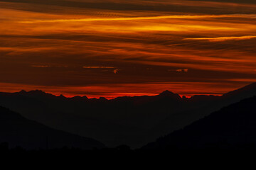 A stunning fiery sunset paints the mountain silhouettes with deep red and orange hues, creating a dramatic and peaceful alpine evening scene.