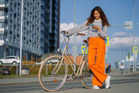 Young woman with a bright smile stands outside in the city with her bicycle, engaging with her smartphone. She represents a blend of urban movement, connectivity, and a laid-back, dynamic city life - Powered by Adobe