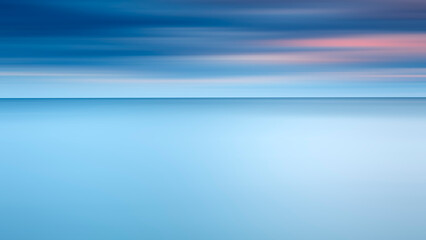 A blurry blue sky and a calm ocean. The sky is covered with blue and red clouds, and the ocean is...