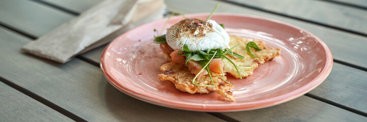 Delicious poached egg on crispy fritter with fresh greens and smoked salmon in pink plate on wooden table.