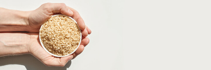 Caucasian male hands holding bowl of uncooked brown rice against white background.