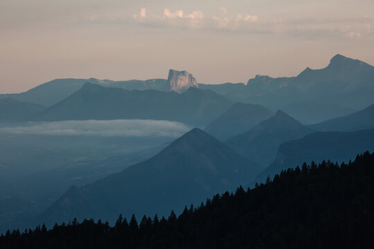 Paysage de montagne au coucher du soleil, avec des couches successives de reliefs bleut&eacute;s, de brume et de for&ecirc;ts sombres. Image apaisante et atmosph&eacute;rique, parfaite pour des projets li&eacute;s &agrave; la nature, 
