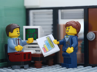 Obraz premium Tambov, Russian Federation - October 19, 2025 A Happy Lego businessperson minifigure sitting behind a computer and talking with another Lego businessperson minifigure that is holding graphs.