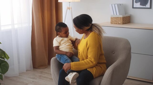 A young African American boy sits on his mother's lap in a comfortable armchair at home. They share a joyful moment, showcasing the warmth of their bond in a bright, inviting room.