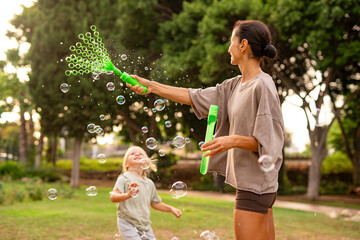 Smiling mother playing with her young son, creating soap bubbles with a wand in the park, joyful family bonding and carefree childhood fun outdoors on a summer day