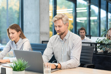 Two call center employees, woman and a man, are working on laptops at their desks while wearing headsets, with colleagues in the background.