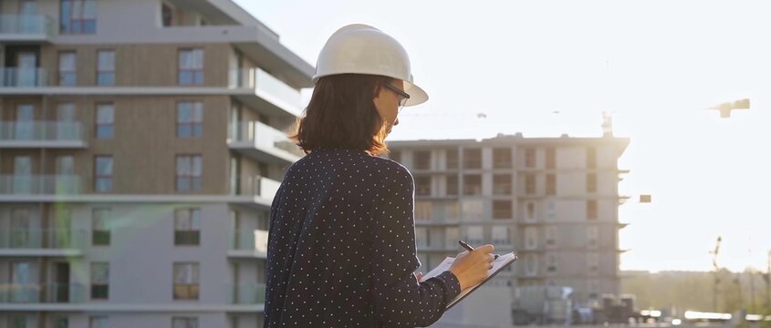 Female architect wearing a safety helmet is using a tablet computer while inspecting a building construction site at sunset
