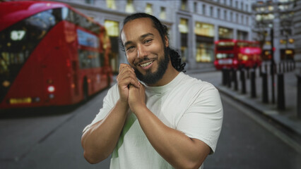 Man clasping hands close to face and smiling on busy city street next to a red doubledecker bus;...
