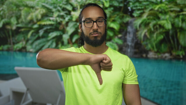 Man giving thumbs down gesture with left thumb at resort pool in neon green shirt and glasses; disapproval criticism.
