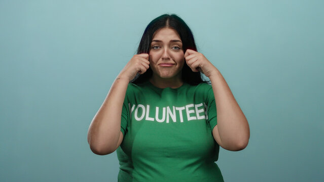 Young woman with a curvy figure, wearing a green volunteer shirt, makes a crying gesture against an isolated blue background wall.