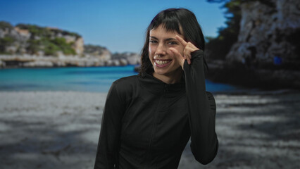 Young hispanic woman in black sportwear pinches fingers street by clear blue sea and rocky cliffs; precision confidence.