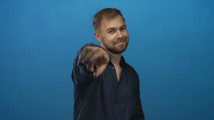 Young man in navy shirt points finger at camera in studio with blue backdrop, smiling and making eye contact; confidence.