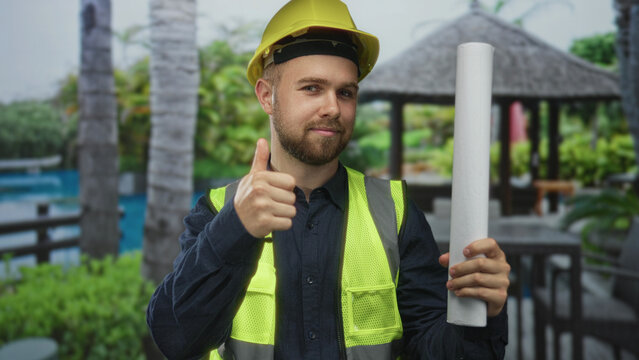 Man construction worker wearing yellow hardhat and high vis vest holding a rolled blueprint and giving thumbs up at building; confidence safety.