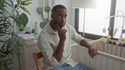 Man with hand on chin sitting beside baby crib in building, lamp and mobile visible, wearing ring on left hand; quiet contemplation.