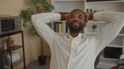 Man with glasses and braces, hands behind head smiling at desk in office wearing white shirt;...