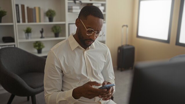 Man using smartphone and gesturing with hands in office building, seated near suitcase and bookshelf; frustration remote work.
