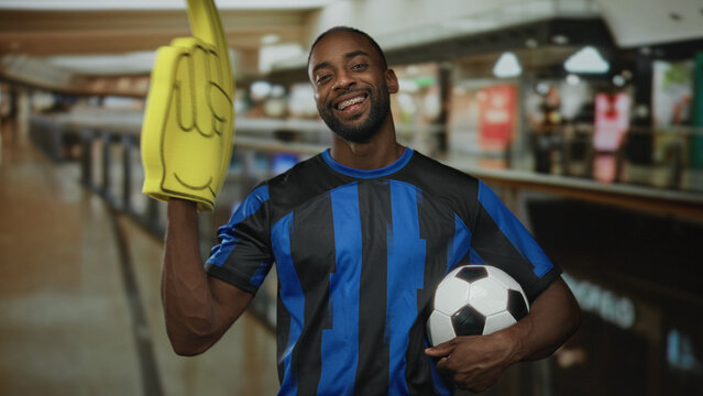 Man holding soccer ball and pointing foam finger upward in building mall corridor while wearing blue striped jersey and braces; team pride.